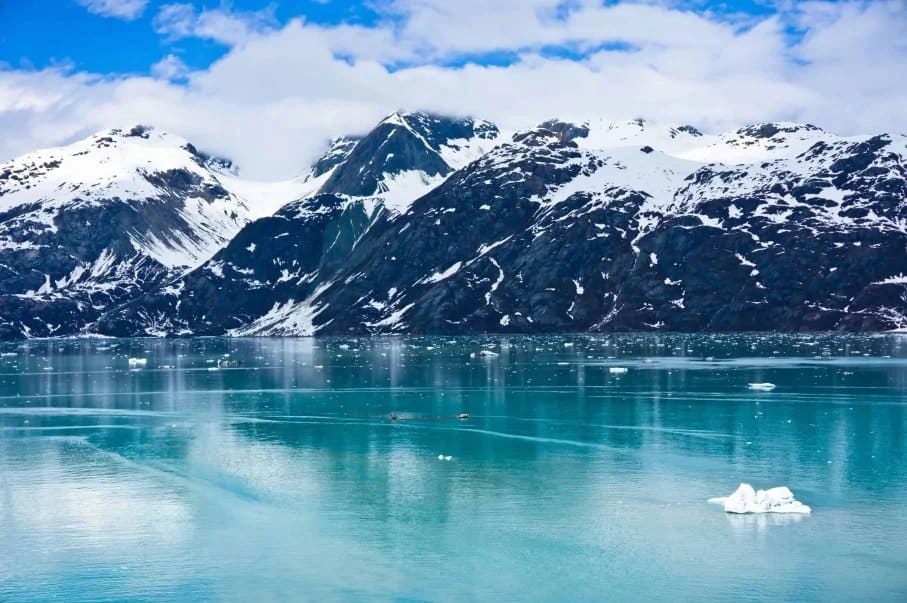 Snow-covered mountains reflected in a turquoise glacial lake under a partly cloudy sky.