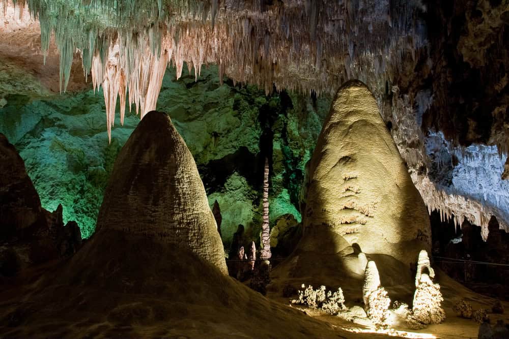 Large stalagmites and stalactites in an illuminated cave chamber with textured rock formations and a greenish background, as highlighted in the Carlsbad Caverns vacation guide.